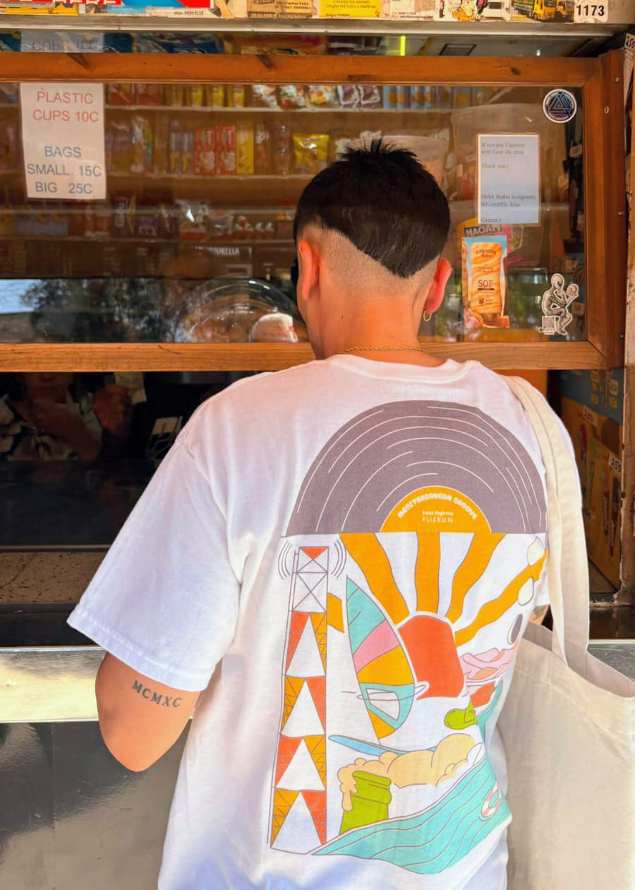 A man viewed from behind wearing a white t-shirt with a large, colorful retro graphic print on the back, standing in front of a vintage-style wooden display counter filled with retro merchandise.
